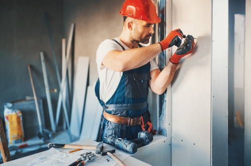 Closeup front view of construction worker placing a plaster board over a wall and using electric screwdriver to tighten it. The worker is wearing blue uniform, red protective gloves and helmet. He's partially unrecognizable with brown beard and mustache.